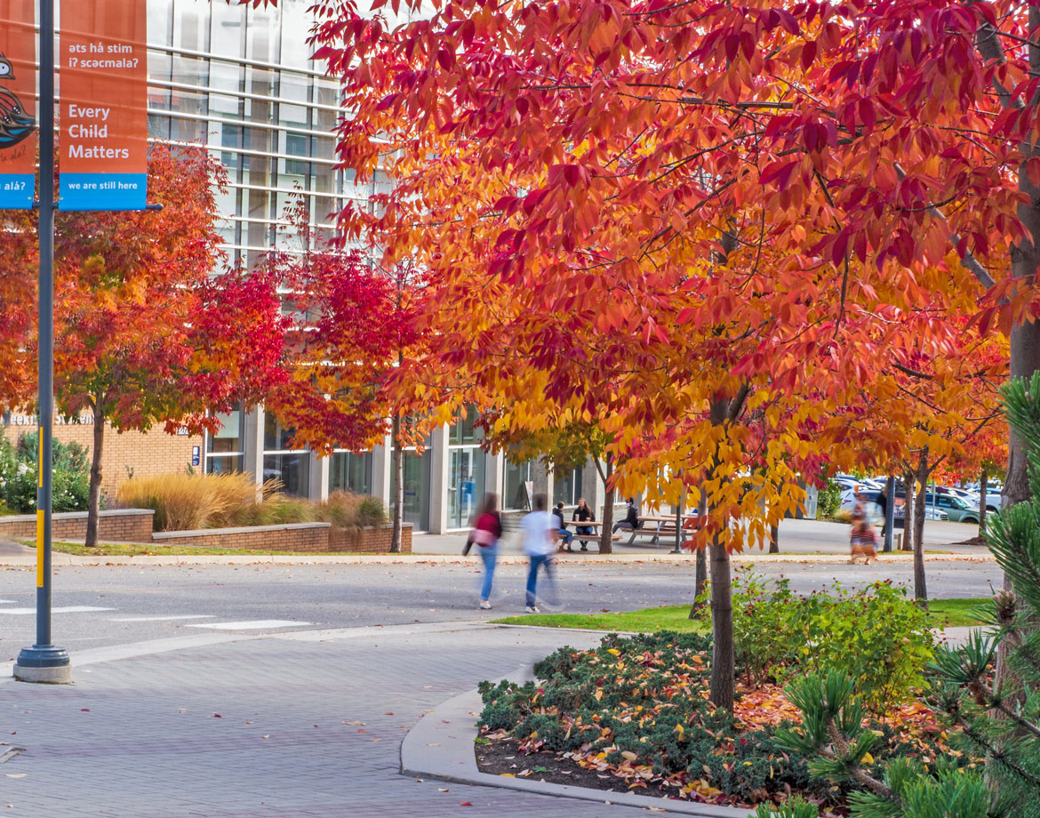 Trees with red and orange leaves in front of UBCO's UNC building, with Every Child Matters banner in midground.