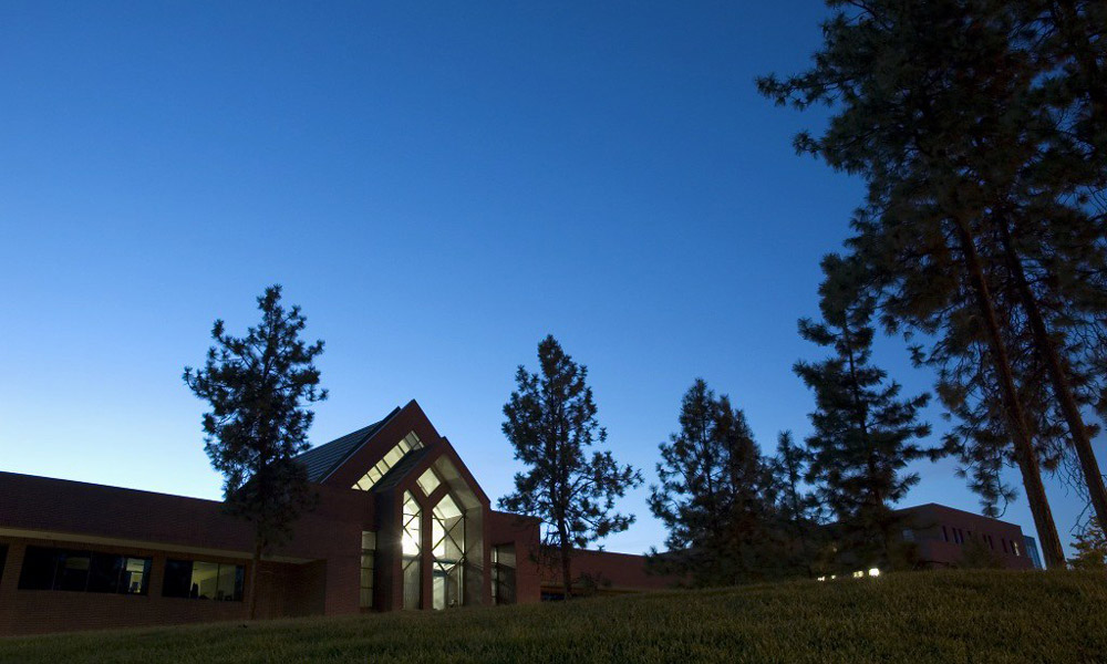 Administration building at UBCO in dark blue dusk, lit from inside with dark silhouettes of trees in front. 