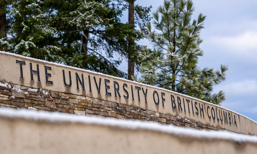 Rock wall with "University of British Columbia" and light snow, pine trees in background. 
