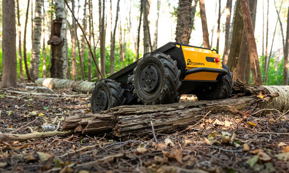 A yellow, ground-based, four-wheel robotic vehicle—Clearpath Robotics’ Husky A300—rolls over forested ground.