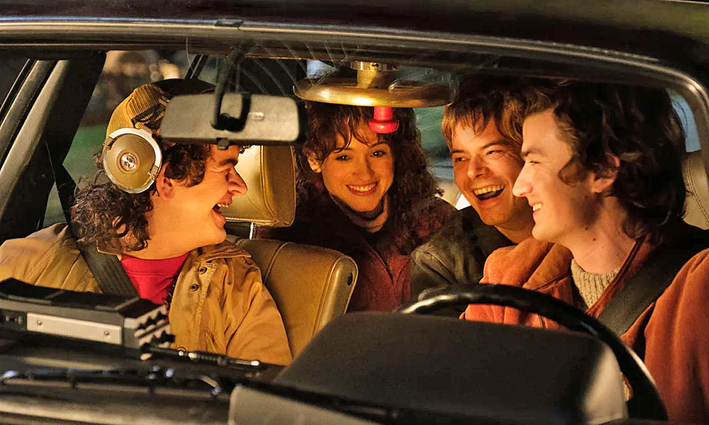 Still image from Stranger Things showing characters Dustin, Nancy, Steve, and Jonathan sitting together in a car at night, laughing and enjoying each other’s company.