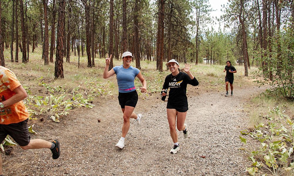 Two young women wave and smile at the camera as they run past during a class celebration. They're in a forest with clearly marked paths.