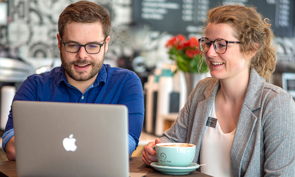 Two UBC medical graduates, Brian Hayes and Hannah Young, seated at a table in a café, smiling while working together on a laptop.
